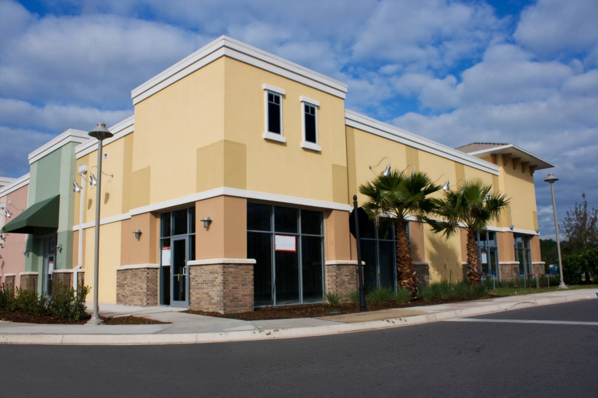 commercial-paving_nj_fhpwnc A vacant yellow commercial building with large windows and palm trees, on a corner under a partly cloudy sky.