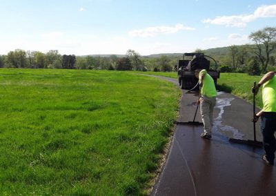 Two workers in yellow shirts pave a winding road through a grassy field under a bright, sunny sky.