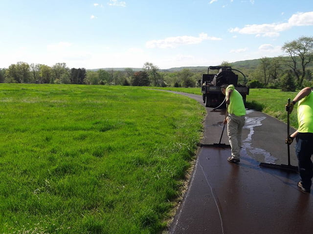 Two workers smooth fresh asphalt on a winding road through a green field, with a truck nearby under clear skies.