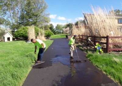 Two workers in yellow shirts are sealing a driveway with tools on a sunny day near a house and tall grass.