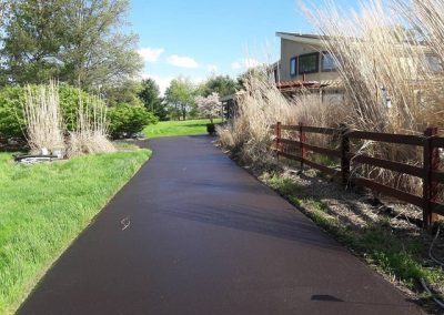 A paved path curves past tall grass, a wooden fence, and a house under a sunny blue sky.