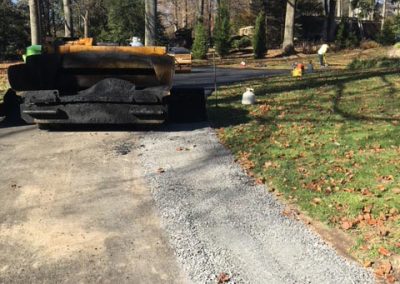 A steamroller and gravel on a driveway under construction, with trees and grass in the background.
