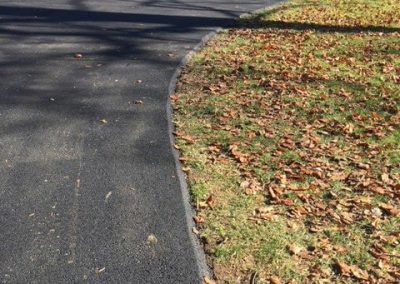 A newly paved winding path bordered by autumn leaves and grass in a wooded area.