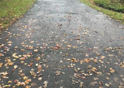 Freshly paved driveway with scattered autumn leaves, leading to a house; a worker and machine in the background.