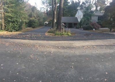 A paved driveway with scattered leaves leads to a house with a carport, surrounded by trees.