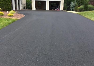 Freshly paved black asphalt driveway leading to a two-car garage in front of a house with greenery around.