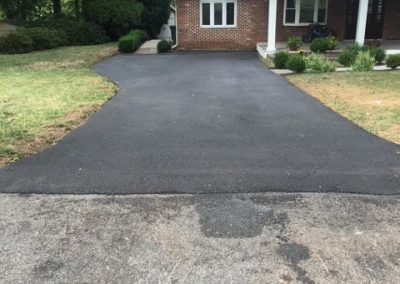 A newly paved black asphalt driveway leads to a brick house with a front porch and green yard.