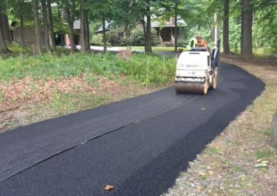 A worker drives a steamroller, paving a fresh asphalt path through a wooded area near houses.
