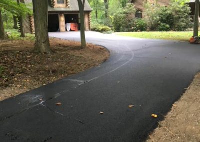 Freshly paved black asphalt driveway winding through trees to a house with a garage in a wooded area.