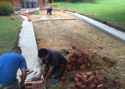 Two people lay bricks for a driveway, with tools and materials next to a house surrounded by greenery.