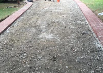 Two workers prepare a gravel driveway bordered by red bricks in front of a house with a porch.