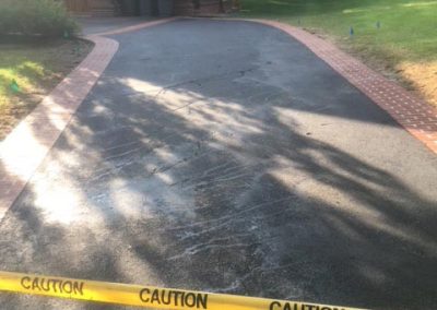 Newly paved driveway with caution tape blocking entry in front of a house surrounded by trees and grass.