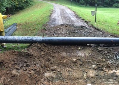 A dirt road blocked by a large pipe and pile of soil, with trees and grass on both sides.