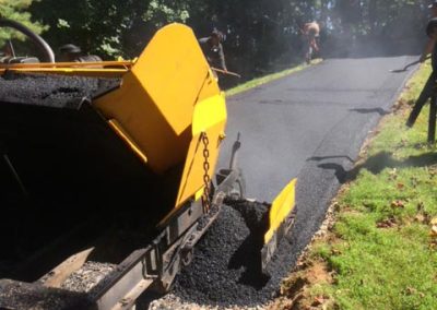 Yellow asphalt paving machine laying fresh black asphalt on a driveway surrounded by trees and grass.