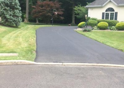 A paved driveway leads to a house with a manicured lawn, shrubs, and trees in the background.