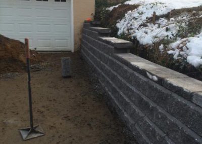 Gray stone retaining wall beside a garage, with snow on the ground and a shovel standing upright.
