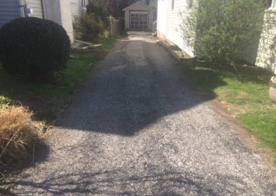 A narrow gravel driveway leads to a small detached garage between two houses on a sunny day.