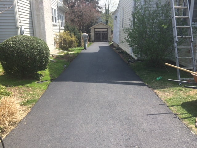 Freshly paved driveway between two houses leading to a small garage; person working in the distance.