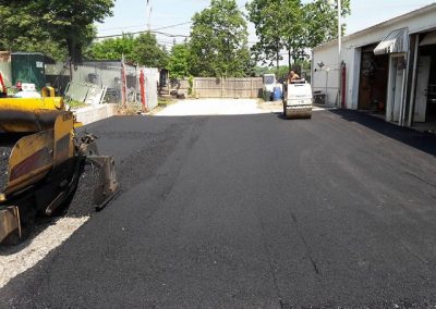 Fresh asphalt being paved outside a garage, with construction vehicles working on the surface.
