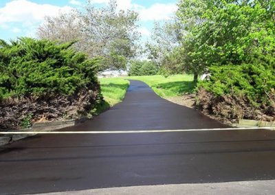 Freshly paved driveway with a yellow barrier across, surrounded by green trees and bushes.