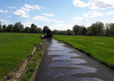 A road in a grassy area is being resurfaced with fresh asphalt by workers under a partly cloudy sky.