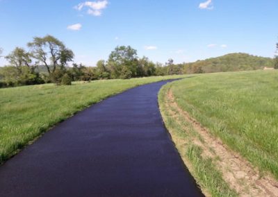 A freshly paved path curves through a grassy field under a blue sky with scattered clouds.