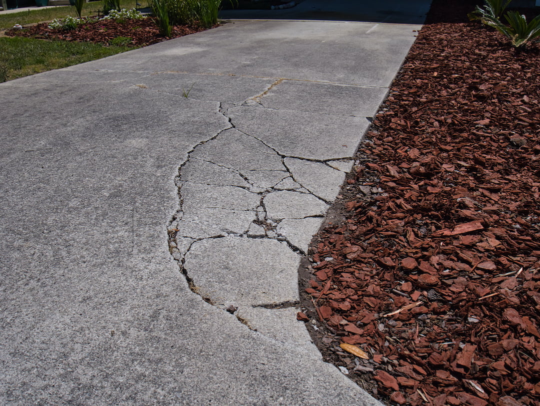 A concrete driveway with large cracks next to red mulch landscaping on a sunny day.