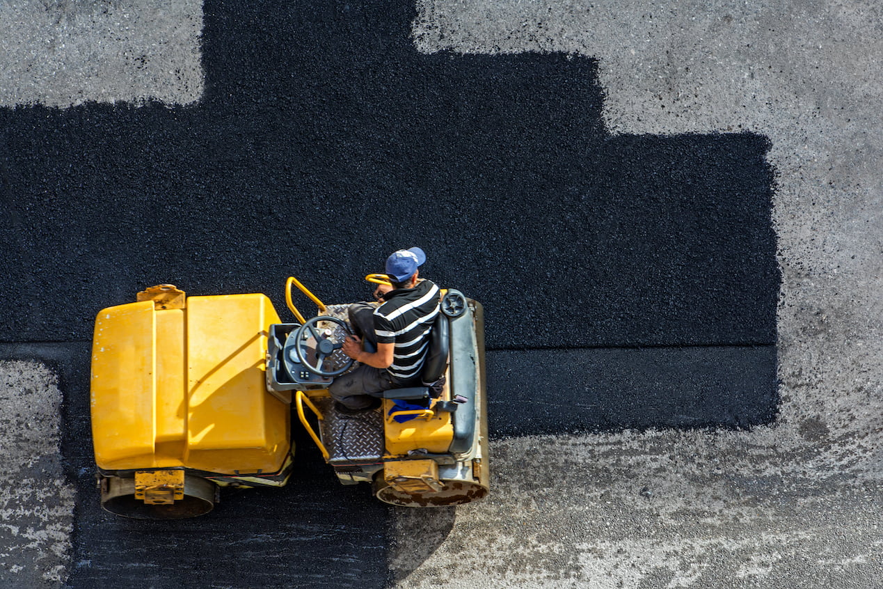Worker operating a yellow road roller compacts fresh asphalt on a road, viewed from above.