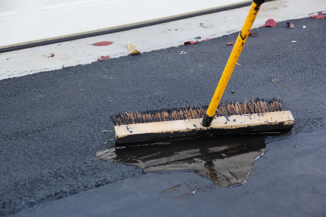 A broom sweeps water from an asphalt driveway near a garage door with a few fallen leaves nearby.