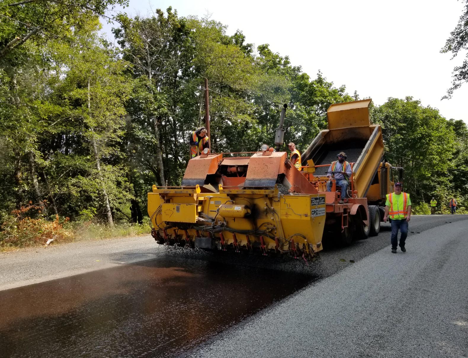 Road crew paving a rural road with heavy machinery, trees and greenery in the background.