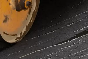 Close-up of a yellow steamroller wheel compacting fresh black asphalt on a road surface.