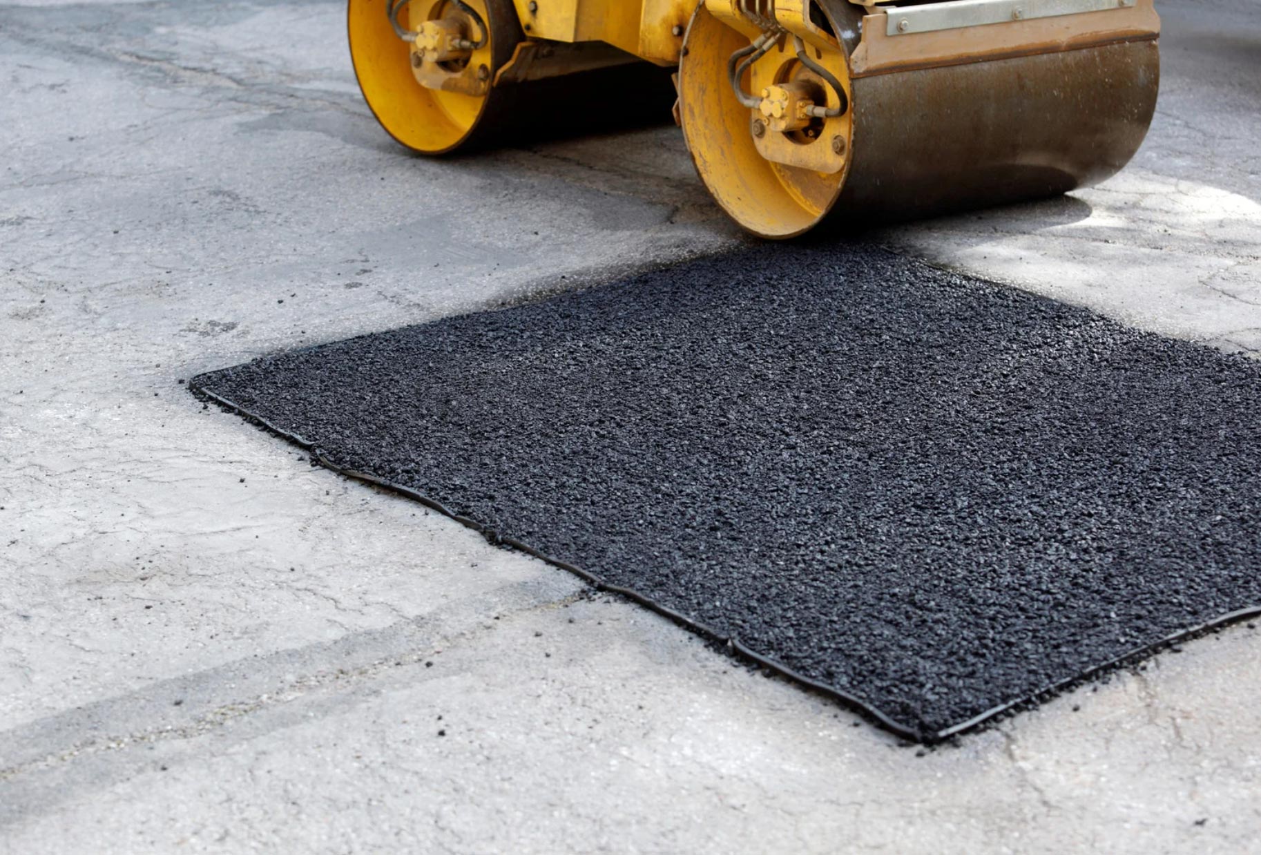 A steamroller compacts fresh asphalt patch on a cracked concrete road surface.