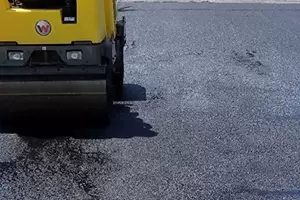 Yellow steamroller compacting fresh black asphalt on a road under bright daylight.
