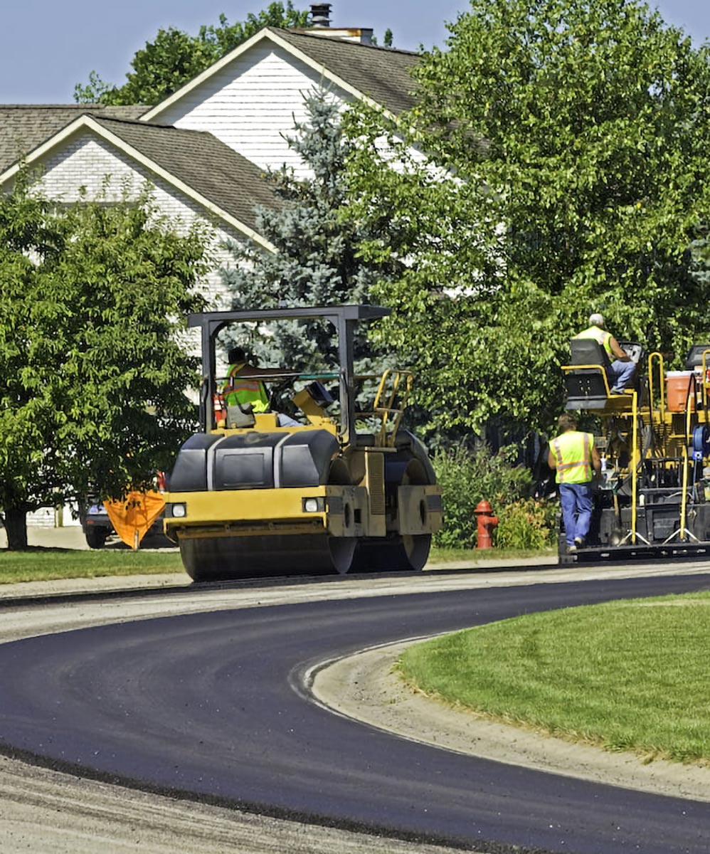 Workers paving a residential street with heavy machinery near houses and green trees on a sunny day.